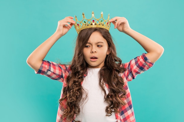 Enfant sérieux aux cheveux bouclés en couronne de princesse sur fond bleu, béat.