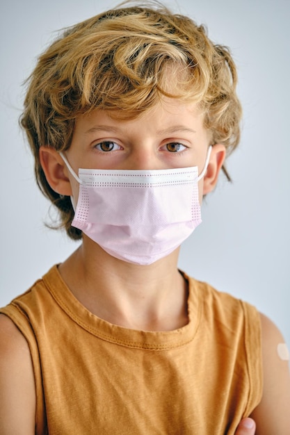 Enfant sérieux aux cheveux bruns portant un masque médical regardant la caméra pendant la pandémie de COVID 19