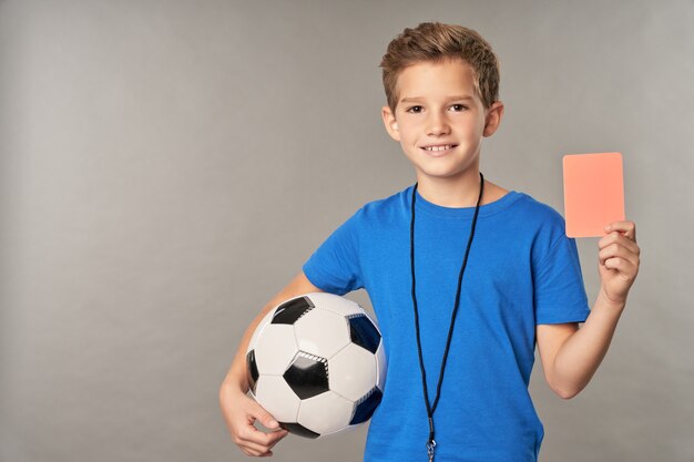 Enfant de sexe masculin mignon en chemise bleue montrant une carte de pénalité et souriant en se tenant debout sur fond gris