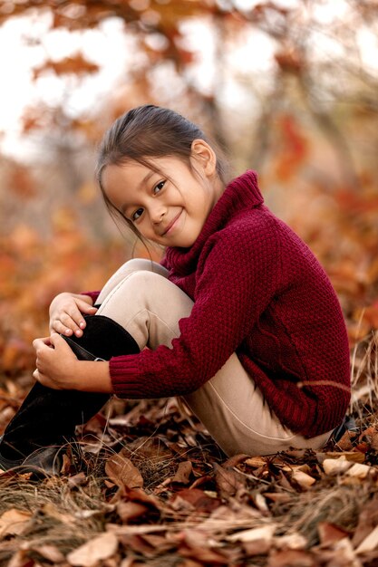 Enfant souriant de 5 ans posant dans le parc en automne. petite fille assise dans les feuilles tombées. enfance heureuse
