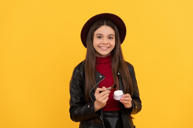 Un enfant souriant au chapeau tient un pinceau de maquillage et de la poudre sur le maquillage de fond jaune