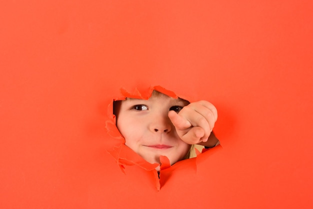 Un enfant souriant jetant un coup d'œil à travers un trou dans un garçon en papier montre le doigt sur vous pour annoncer des produits pour enfants