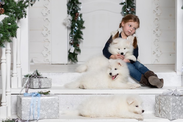 Un enfant souriant mignon embrasse un chien de race Samoyède blanc. Une fille blonde, a un visage heureux, de jolis yeux, embrasse un chien husky samoyède. Maison de vacances portrait. Animal de famille d'hiver. Style de mode pour enfants.