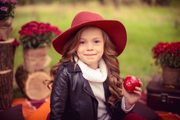 Enfant souriant avec panier de pommes rouges, assis dans un parc en automne