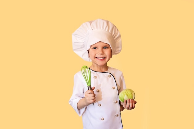 Enfant souriant en uniforme de chef blanc, tenant un chou et un fouet dans ses mains. Joyeux fond jaune.