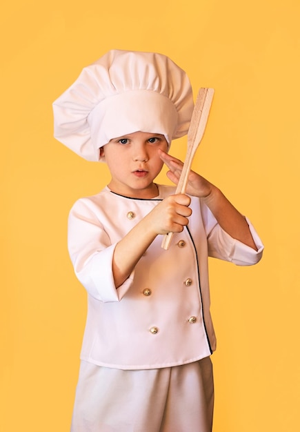 Enfant souriant en uniforme de chef blanc tenant une spatule en bois dans les mains Fond jaune joyeux