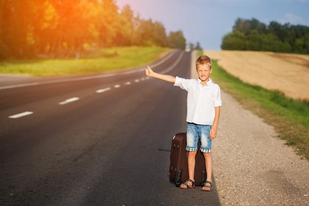 Enfant souriant avec valise voyageant en stop. route d&#39;été