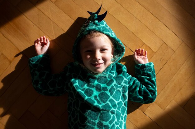 Une enfant souriante en costume de dinosaure est allongée sur le sol.