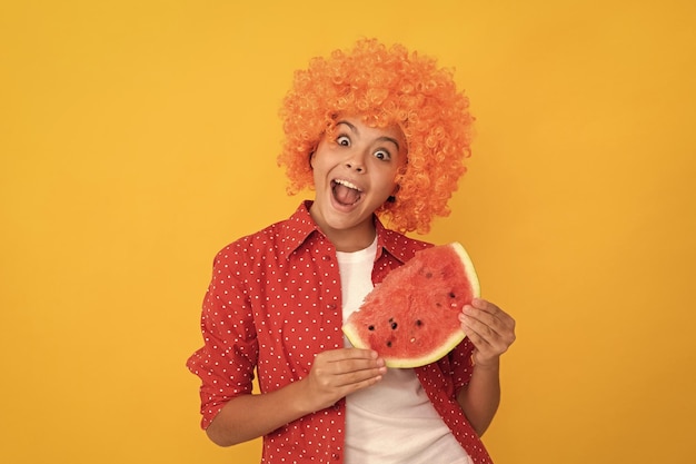 Enfant surpris dans une perruque de cheveux orange fantaisie tenant une tranche de melon d'eau fraîche et mûre