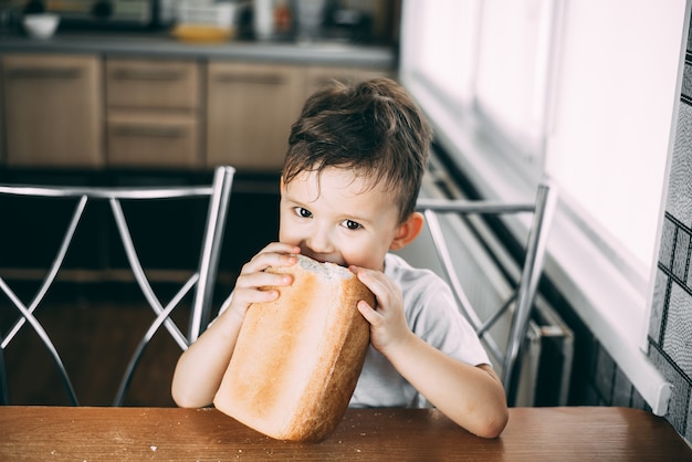 L'enfant à table pendant la journée mange avidement une miche de pain entière en forme de brique