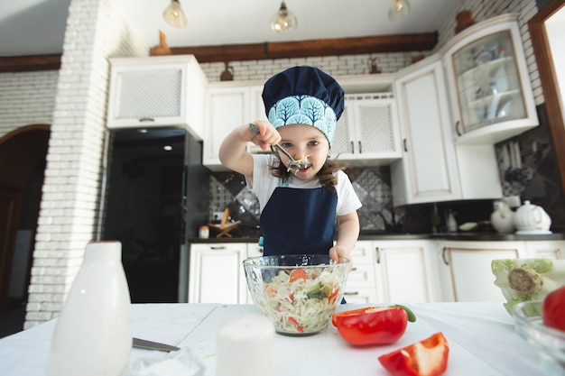 Un enfant en tablier et toque mange une salade de légumes dans la cuisine