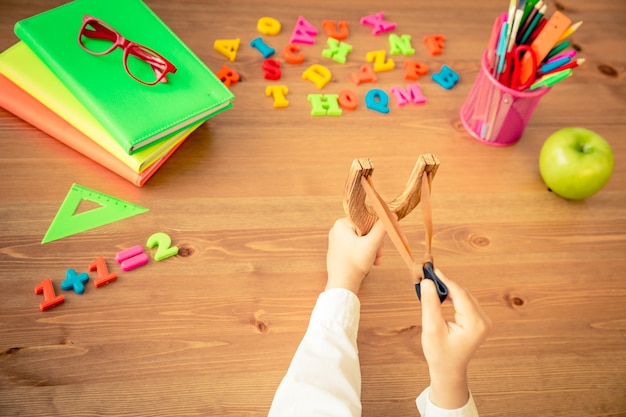 Enfant tenant une fronde dans les mains. Articles scolaires sur un bureau en bois en classe. Notion d'éducation. Vue de dessus