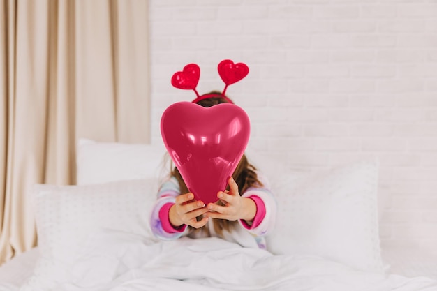 L'enfant tient un ballon en forme de coeur une petite fille est allongée sur le lit et tient un coeurSaint Valentin