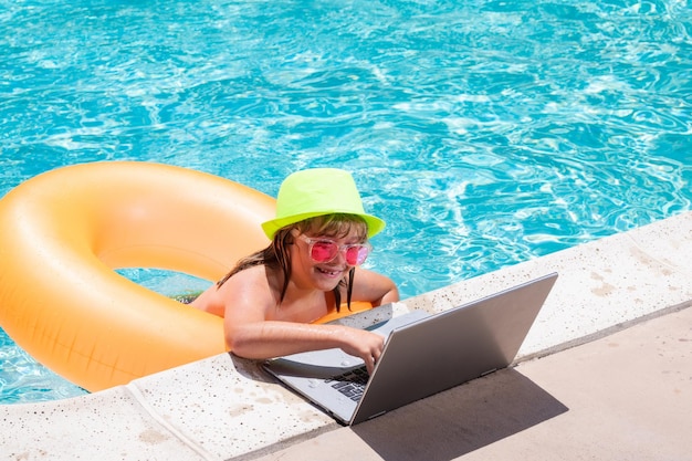 Enfant travaillant à distance sur un ordinateur portable dans la piscine Petit homme d'affaires travaillant en ligne sur un ordinateur portable dans l'eau de la piscine d'été Petit homme d'affaires travaillant sur une plage tropicale