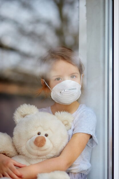 Enfant triste, petite fille au masque avec ours en peluche à la recherche de fenêtre, coronavirus en quarantaine