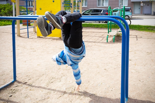 Enfant en veste bleue accrochée à des barres transversales à l'envers dans une aire de jeux, sur fond de maison et de voitures.