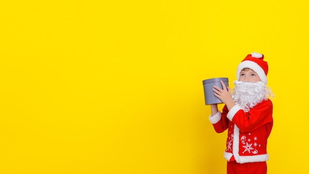 Un enfant en vêtements de père Noël et avec une barbe artificielle tient une boîte-cadeau ronde dans ses mains