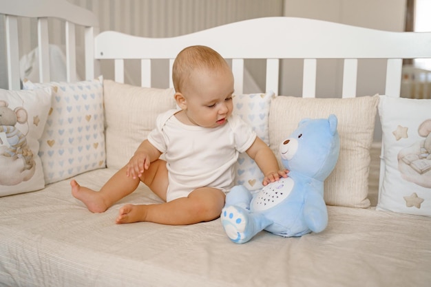 Un enfant vêtu d'un body blanc dans un berceau blanc est assis sur son cul à côté d'un ours en peluche