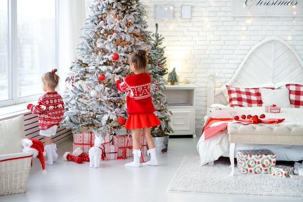 Les enfants accrochent des jouets et des boules de Noël sur un arbre de Noël artificiel dans une chambre blanche