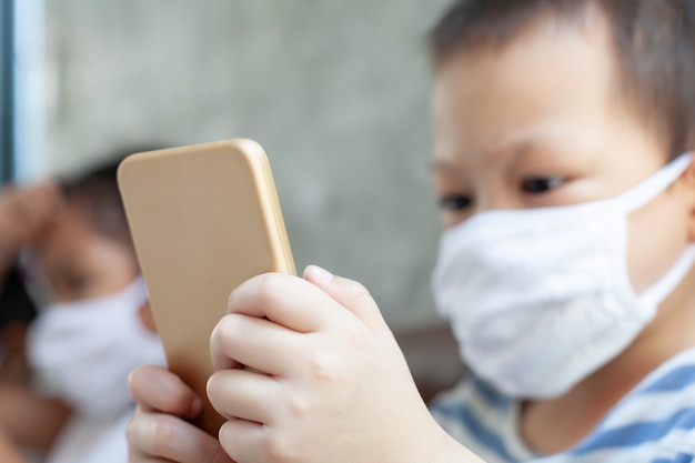 Enfants asiatiques garçon et filles portant un masque de protection jouant sur le smartphone ensemble à la quarantaine à domicile contre le coronavirus Covid-19 et la pollution de l'air pm2.5.