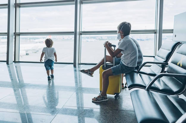 Enfants au terminal de l'aéroport vide attendant le départ en regardant par la fenêtre. Des petits garçons portant un masque médical protecteur sont assis dans le salon en attendant le vol d'avion. Concept de voyage en famille