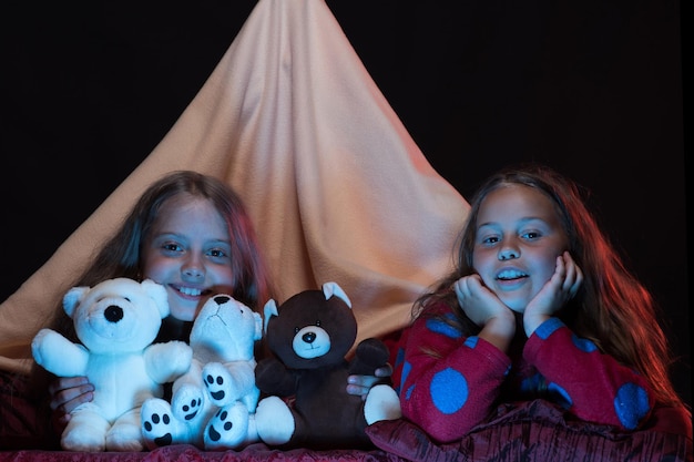 Enfants et concept de temps amusant PJs party pour enfants Filles avec des visages heureux jouant avec des nounours Enfants portant des pyjamas rouges au lit sur fond noir Amis s'amusant avec des jouets dans une tente de couverture