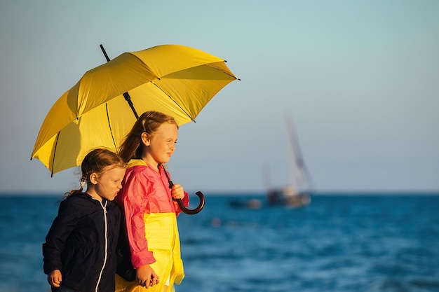 enfants dans des vêtements imperméables brillants et avec un porte-parapluie jaune au bord de la mer