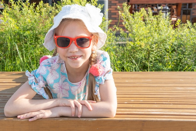 Enfants et été Une petite fille heureuse en lunettes de soleil et un chapeau rit et sourit à la caméra allongée sur un banc