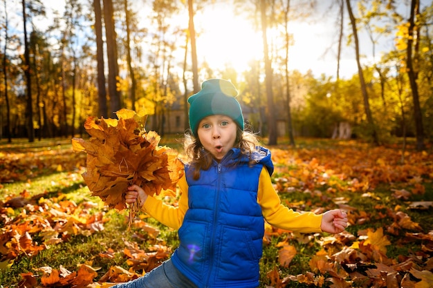 Enfants à l'extérieur Drôle adorable petite fille recueille des feuilles d'érable sèches tombées jouant parmi les feuilles jaunes dans un parc d'automne avec des rayons de soleil tombant au coucher du soleil Concept d'une enfance heureuse et insouciante