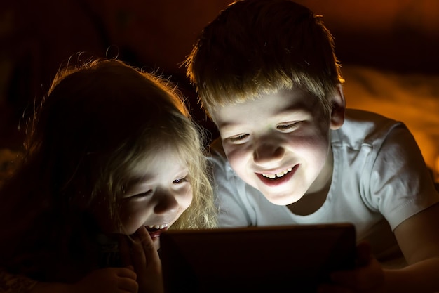 Les enfants garçon et fille allongés sur le lit sourient regarder des dessins animés sur un ordinateur portable la nuit avant le coucher