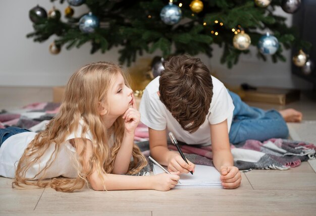 Enfants - un garçon et une fille écrivent des lettres au Père Noël près de l'arbre de Noël dans le salon