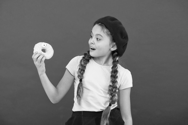 Enfants grands fans de beignets cuits au four Impossible de résister à un beignet frais Une fille tient un joli beignet glacé à la main sur fond rouge Enfant fille souriante prête à manger un beignet Boutique de bonbons et concept de boulangerie