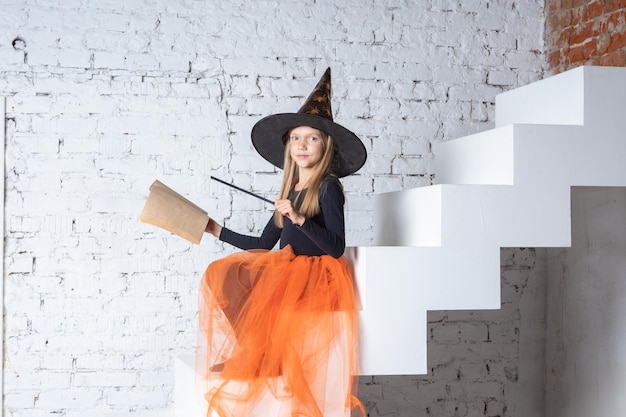 Enfants Halloween Une belle fille douce dans un costume de sorcière portant un chapeau tenant un livre avec des sorts et une baguette magique