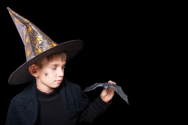 Enfants D'halloween. Un Garçon Dans Un Chapeau De Sorcier Et Avec Une Araignée Peinte Sur Sa Joue Tenant Une Chauve-souris Noire D'halloween Pour Effrayer Quelqu'un Isolé Sur Un Fond Noir. Prêt Pour Le Tour De Passe-passe Des Fêtes.