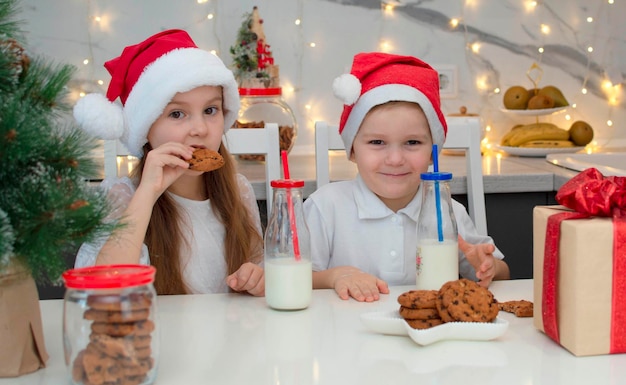 Des enfants heureux en bonnets de Noel se racontent des histoires drôles. Sœur et frère boivent du lait, mangent des biscuits près de la maison dans la cuisine avec une décoration de Noël la veille de Noël à la maison. Mise au point sélective.