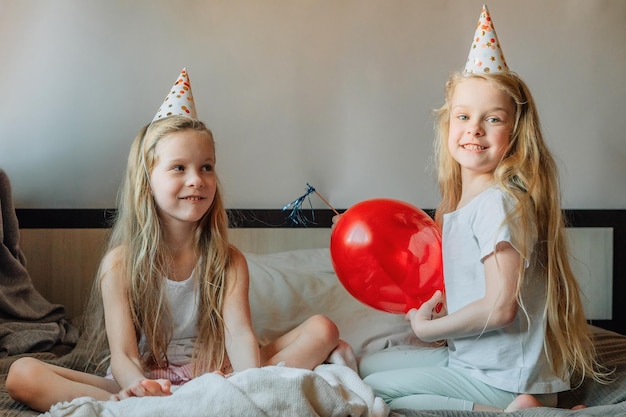 Enfants heureux filles soeurs jumelles à la maison sur le lit le matin le jour de leur anniversaire en casquettes festives avec un ballon rouge en forme de coeur