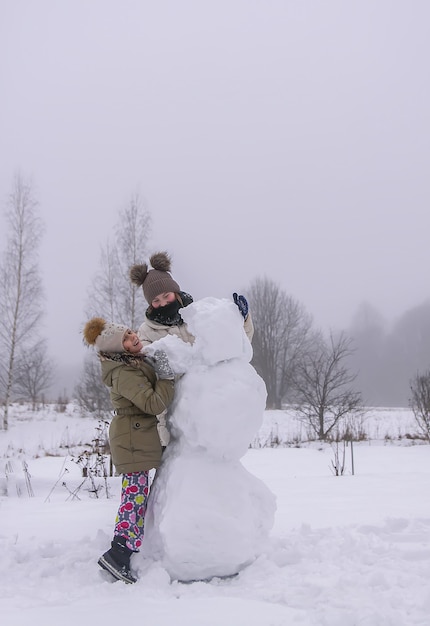 Des enfants heureux font un bonhomme de neige dans un champ enneigé à la campagne.