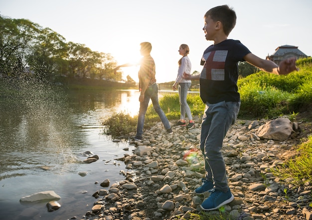 Photo enfants heureux jouant près de la rivière. garçons et filles jetant des cailloux dans la rivière. journée ensoleillée d'été