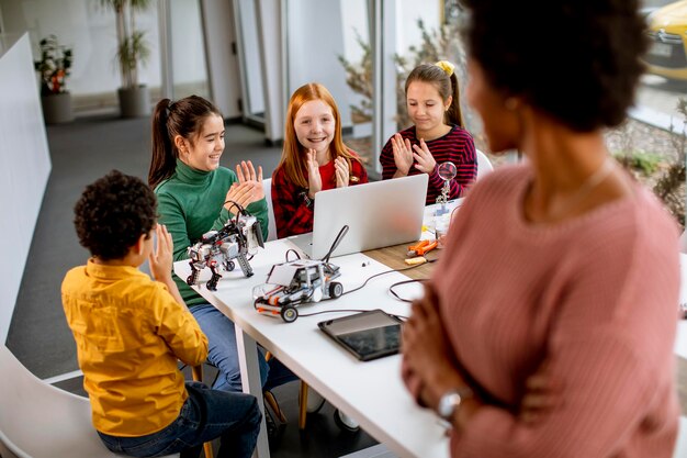 Des enfants heureux avec leur professeur de sciences afro-américaine avec un ordinateur portable programmant des jouets électriques et des robots en classe de robotique