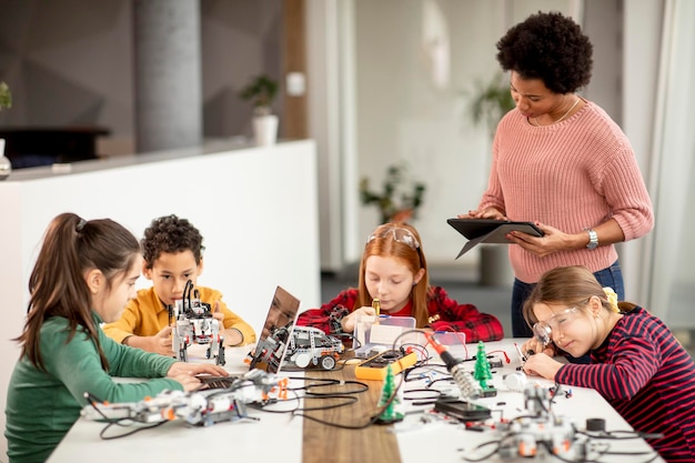 Des enfants heureux avec leur professeur de sciences afro-américaine avec un ordinateur portable programmant des jouets électriques et des robots en classe de robotique