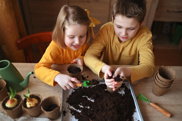 Photo des enfants heureux plantent des plantes à la maison, une fille et un garçon sèment des graines dans des pots de jardinage naturels