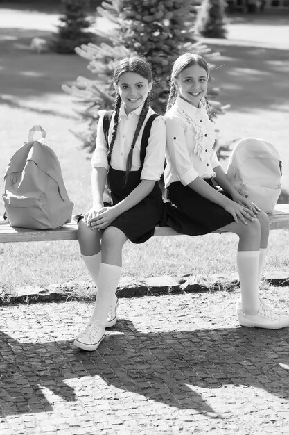 Des enfants heureux en uniforme formel s'assoient sur un banc de parc avec des sacs se relaxant après la journée d'école à l'extérieur des élèves