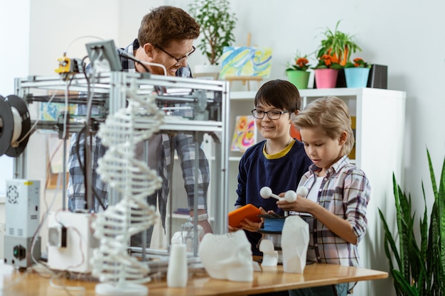 Enfants intéressés inspectant l'imprimante 3D placée sur la table de travail et touchant des exemples de son fonctionnement