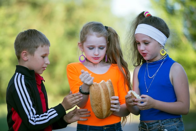 Des enfants joyeux, un garçon et deux filles vêtus de vêtements brillants mangent un petit pain dans la rue