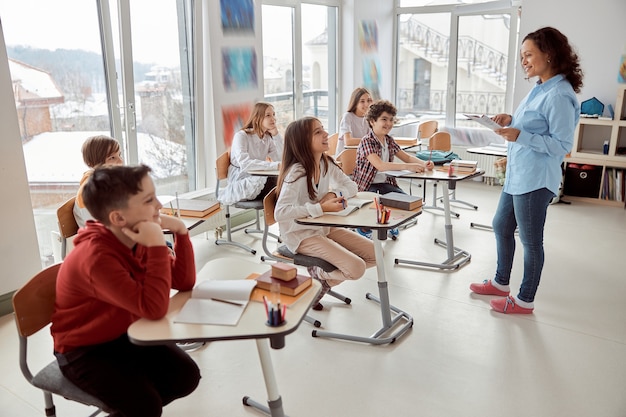 Enfants joyeux et heureux assis au bureau pendant que l'enseignant parle dans la classe de l'école. Enfants de l'école élémentaire assis sur un bureau.