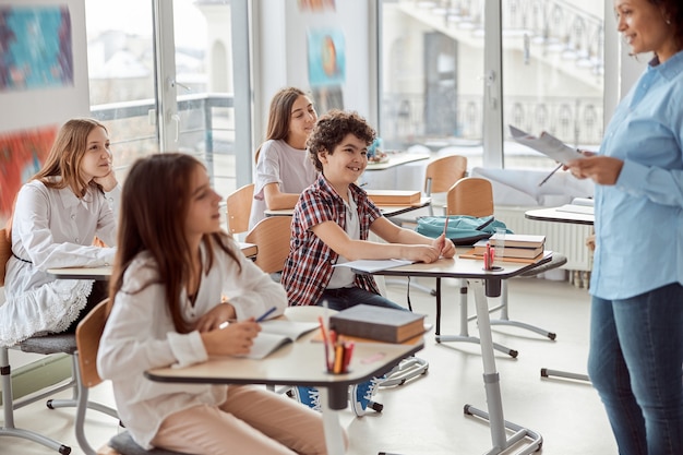 Enfants Joyeux Et Heureux Assis Au Bureau Pendant Que L'enseignant Parle Dans La Classe De L'école. Enfants De L'école élémentaire Assis Sur Un Bureau.