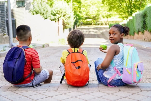 Enfants multiethniques avec des sacs à dos assis dans la rue à l'entrée de l'école mangeant des pommes