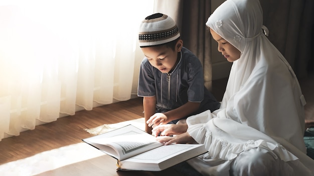 Les enfants musulmans asiatiques religieux apprennent le Coran et étudient l'islam après avoir prié Dieu à la maison. La lumière du coucher du soleil brille à travers la fenêtre. Climat chaud paisible et merveilleux.