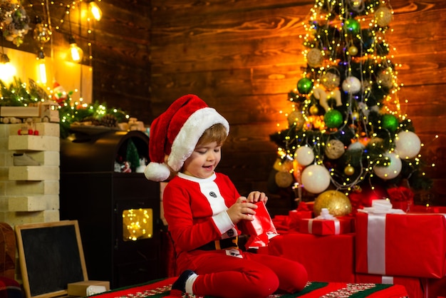 Enfants de Noël. Heureux petit enfant porte des vêtements de Santa, jouant avec une boîte-cadeau de Noël entourée de cadeaux de Noël.