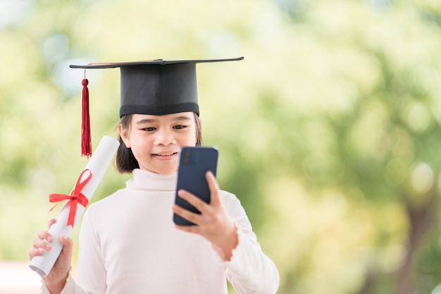 Les enfants obtiennent leur diplôme avec une casquette de remise des diplômes et un certificat de remise des diplômes roulé tenant un smartphone